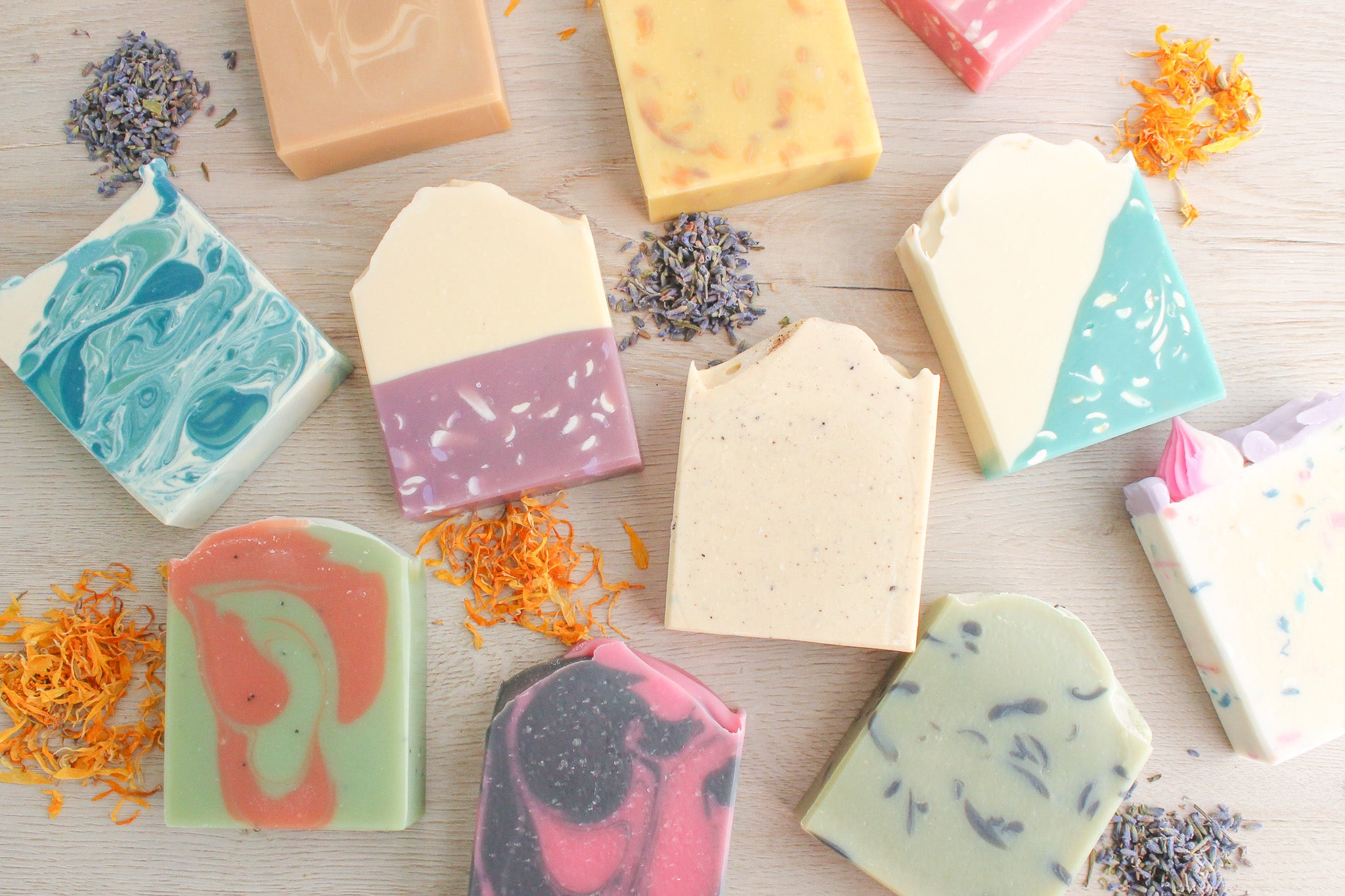 Assorted colorful soap bars on a light wooden surface with lavender and orange flower petals.